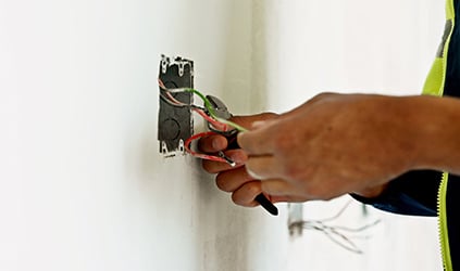 | Patrick Riley Services Close-up of a worker's hand wiring an electrical box in the wall, with exposed red, green, and white wires.