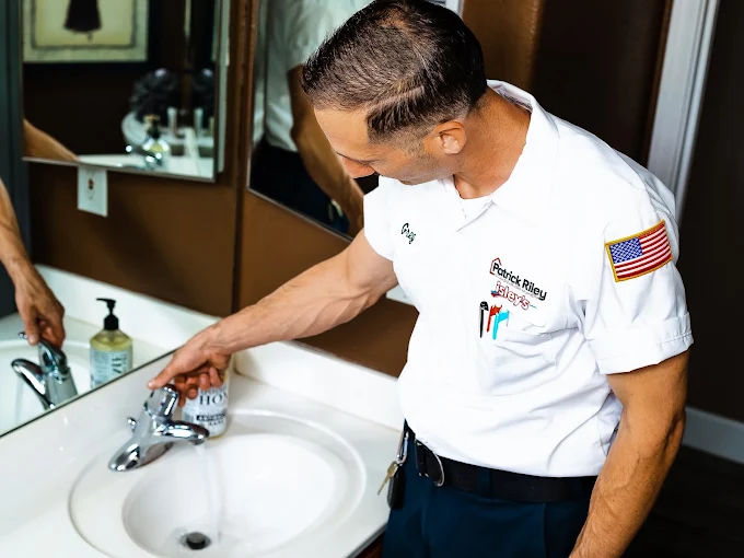 | Patrick Riley Services Plumber in a white uniform with USA patch using a bathroom sink faucet, soap bottle on the counter nearby.